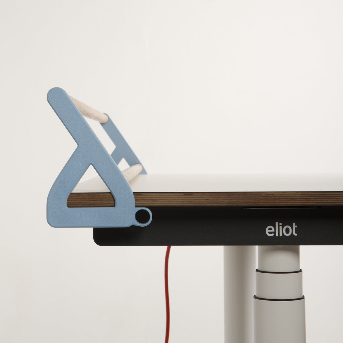 Ein moderner Schreibtisch mit einer blauen, geometrischen Buchstütze auf der linken Seite, einer Tischplatte aus Holz, zylindrischen weißen Metallbeinen, einem sichtbaren roten Kabel und dem Schriftzug „eliot“ auf dem schwarzen Rahmen – perfekt ergänzt durch Schreibtischzubehör wie den „Tiny Top Schreibtischaufsatz Hellblau“ von Smartfurniture GmbH.