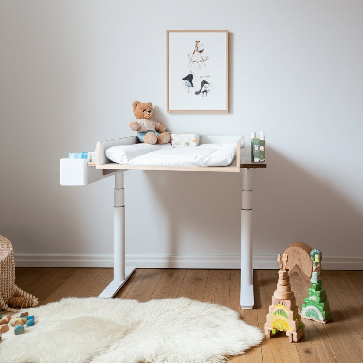In einem gemütlichen Kinderzimmer steht der Wickelaufsatz der Smartfurniture GmbH (für Eliot Tiny White Tischplatte 100x60cm), mit einem Teddybär, Lotionflaschen und einem gerahmten Tiermotiv darüber, sowie Holzspielzeug und einem weichen Teppich auf dem Holzboden.