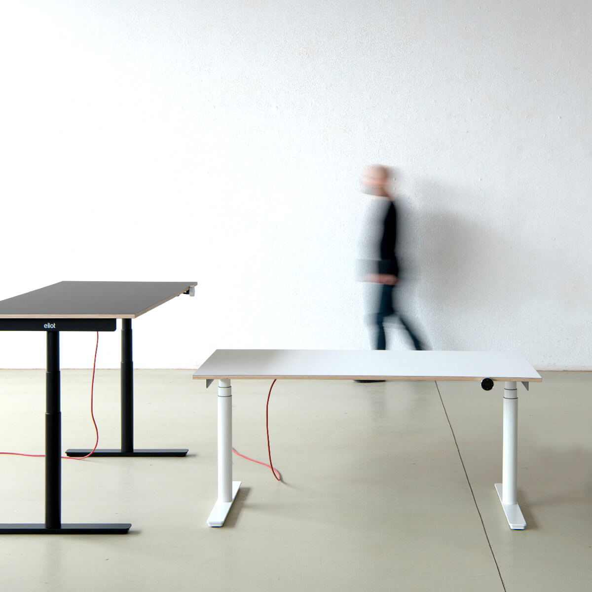 Profil eines schwarzen Eliot-Tisches mit grauer Tischplatte in einem leeren Raum neben einem kleinen Eliot in weiß, im Hintergrund eine verschwommene Person in Bewegung