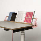 Ein moderner Schreibtisch mit hellgrauer Oberfläche besticht durch eine hervorragende Schreibtischorganisation, unter anderem dank des „Tiny Top“-Schreibtischaufsatzes in Rosa von Smartfurniture GmbH – das ideale 3D-Druck-Zubehör für einen aufgeräumten Arbeitsplatz. Auf dem rosa Organizer liegen ein Tablet, ein blaues Buch und ein aufgeschlagenes Buch mit dem Text „Its APEROL oclock“. Das weiße Tischbein trägt die Aufschrift „eliot“ und rundet diese stilvolle Einrichtung perfekt ab.