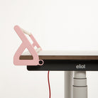 Ein stilvoller, rosafarbener „Tiny Top Schreibtischaufsatz Rosa“ von Smartfurniture GmbH sorgt für moderne Ordnung am Rand eines Holzschreibtisches namens „Eliot“, hält Bücher aufrecht und optimiert gleichzeitig den Arbeitsbereich. Unter dem Schreibtisch hängt ein rotes Kabel, das sich vor einem schlichten weißen Hintergrund abhebt.