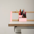 Ein minimalistischer Schreibtisch mit dem „Tiny Top Kids Set Schreibtischaufsatz Rosa“ von Smartfurniture GmbH – einem rosa Aufbewahrungselement für Bleistifte, Kugelschreiber und Buntstifte – steht vor einem schlichten, hellen Hintergrund und eignet sich somit perfekt als kreativer Lern- und Maltisch, der die Fantasie der Kleinen beflügelt.