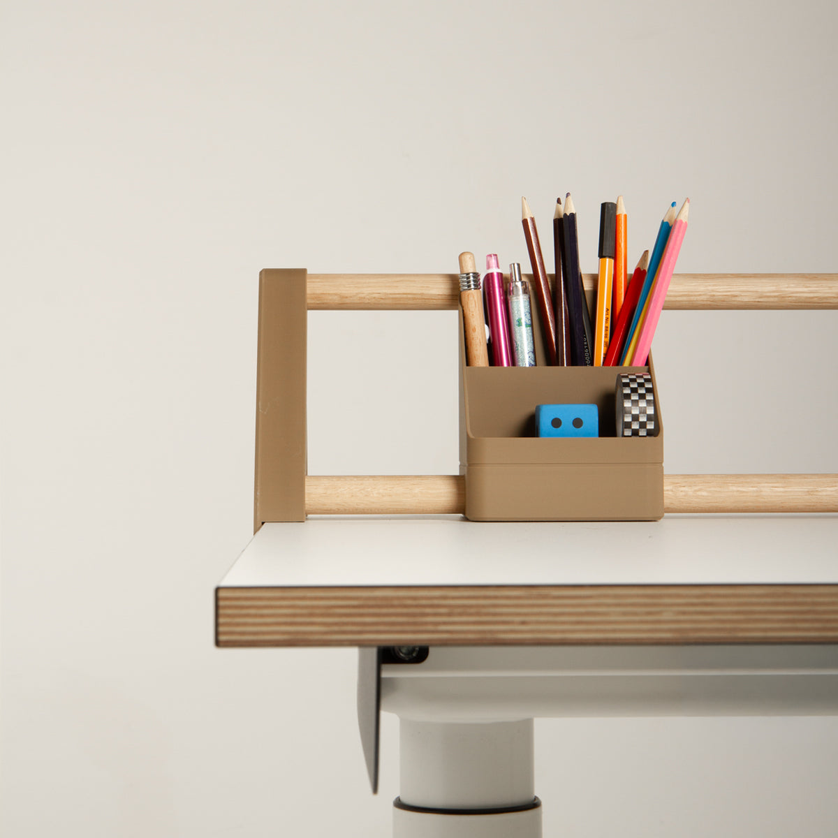Ein beiges „Tiny Top Kids Set Schreibtischaufsatz Mokka“ von Smartfurniture GmbH, bestehend aus Bleistiften, Kugelschreibern, einem Radiergummi und einem Anspitzer, ist an der Holzleiste eines modernen weißen Schreibtisches mit einer Kante aus hellem Eichenmassivholz befestigt, der vor einem schlichten Hintergrund steht.
