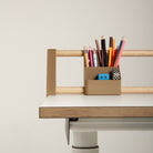Ein beiges „Tiny Top Kids Set Schreibtischaufsatz Mokka“ von Smartfurniture GmbH, bestehend aus Bleistiften, Kugelschreibern, einem Radiergummi und einem Anspitzer, ist an der Holzleiste eines modernen weißen Schreibtisches mit einer Kante aus hellem Eichenmassivholz befestigt, der vor einem schlichten Hintergrund steht.