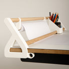 Das „Tiny Top Kids Set Schreibtischaufsatz White“ von Smartfurniture GmbH mit Holzstangen für eine Rolle weißes Papier und einem Fach für Buntstifte und Kugelschreiber steht am Rand eines kreativen Lern- und Maltisches – perfekt, um die Kreativität an Ihrem Schreibtisch für Kinder anzuregen.