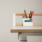 Ein weißes „Tiny Top Kids Set Schreibtischaufsatz White“ von Smartfurniture GmbH – ideal als Schreibtisch für Kinder – verfügt über eine Holz- und weiße Leiste, auf der ein weißer Organizer mit Bleistiften, Kugelschreibern, einem Radiergummi, einem Anspitzer und einem blauen Würfel angebracht ist, alles vor einem schlichten, hellen Hintergrund. Dieser kreative Lern- und Maltisch bietet Kindern einen inspirierenden Raum für Kreativität und Lernen.