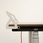 Eine minimalistische Nahaufnahme zeigt einen modernen Schreibtisch mit dem „Tiny Top Schreibtischaufsatz White“ von Smartfurniture GmbH, der elegant an der Kante befestigt ist und so für eine optimale Organisation des Schreibtisches sorgt. Auf dem schwarzen Rahmen des Schreibtisches ist das „eliot“-Logo zu sehen, während darunter ein rotes Kabel herunterhängt. Der Hintergrund ist schlicht weiß.
