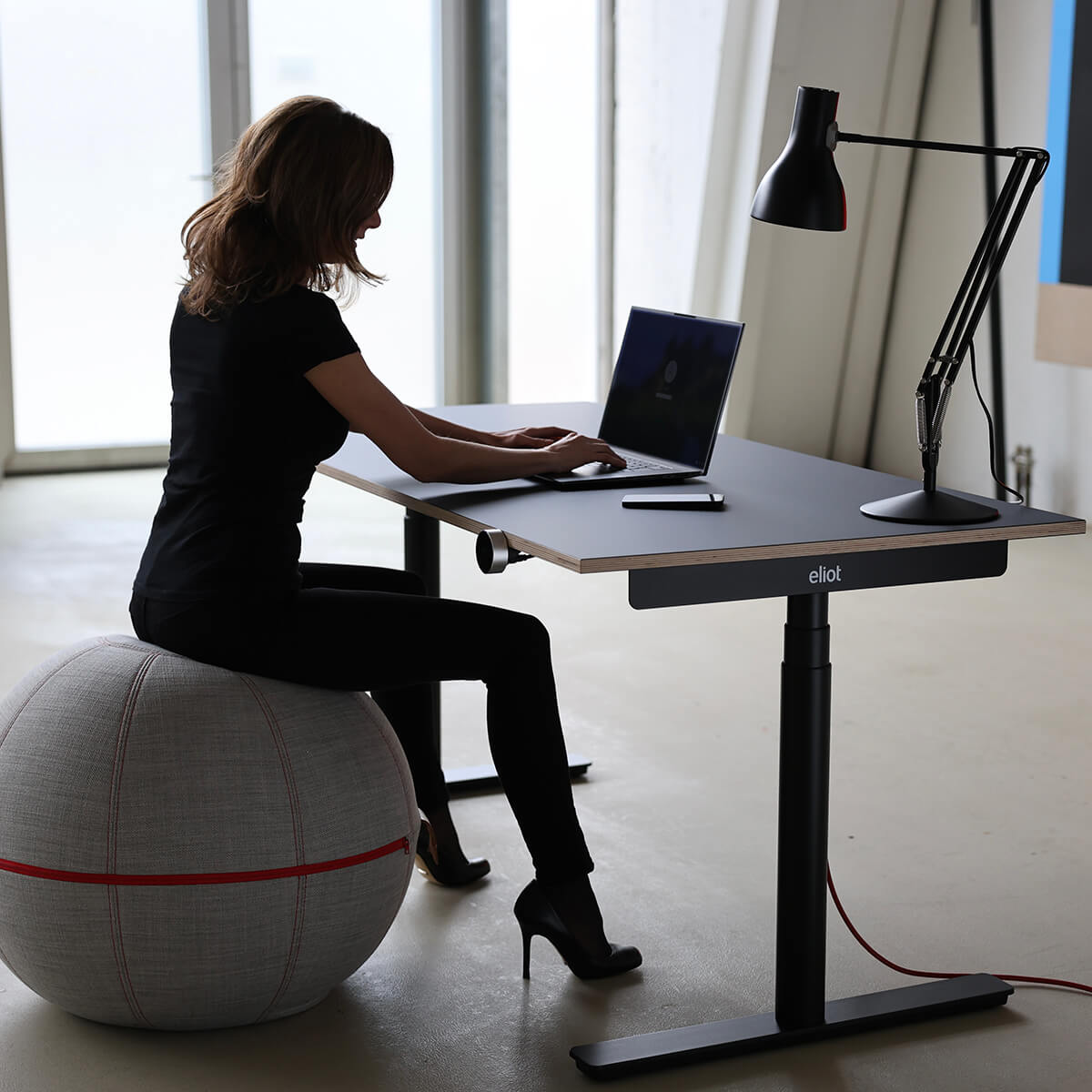 Ein Frau sitzt auf einem großen Stoffball und arbeitet am Laptop, der an auf einem schwarzen Eliot-Tisch liegt
