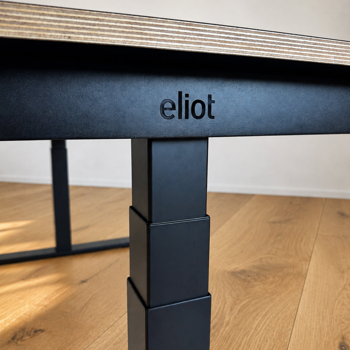 Nahaufnahme des Eliot Pro Black von Smartfurniture GmbH: ein schwarzes Metallbein mit eingraviertem "eliot", das eine 180x80cm große Tischplatte aus Eichenmultiplex trägt. Dieser höhenverstellbare Schreibtisch verfügt über fortschrittliche Doppelmotor-Technologie.