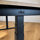 Nahaufnahme des Eliot Pro Black von Smartfurniture GmbH: ein schwarzes Metallbein mit eingraviertem "eliot", das eine 180x80cm große Tischplatte aus Eichenmultiplex trägt. Dieser höhenverstellbare Schreibtisch verfügt über fortschrittliche Doppelmotor-Technologie.