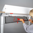 Eine Person legt ein rotes Ladekabel in eine schwarze Gitterschale, die unter einem Wickelaufsatz der Smartfurniture GmbH (für Eliot Tiny White Tischplatte 100x60cm) angebracht ist.