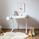 In einem gemütlichen Kinderzimmer steht der Wickelaufsatz der Smartfurniture GmbH (für Eliot Tiny White Tischplatte 100x60cm), mit einem Teddybär, Lotionflaschen und einem gerahmten Tiermotiv darüber, sowie Holzspielzeug und einem weichen Teppich auf dem Holzboden.